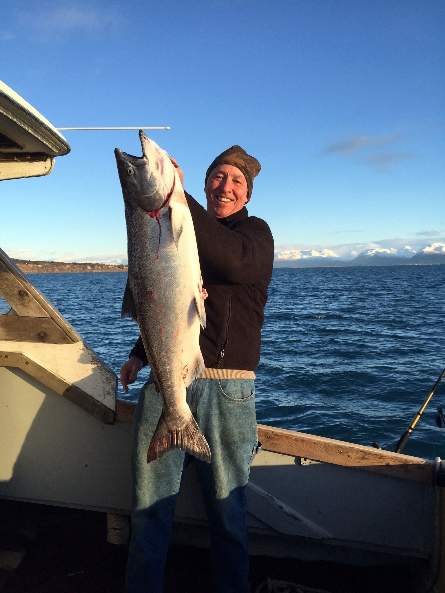 Une personne tenant un gros poisson sur un bateau, avec une vue dégagée sur l’eau et les montagnes au loin.