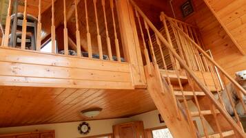 Interior de una cabaña de madera con una escalera que conecta dos pisos, una zona de cocina y un ventanal con vistas al exterior.