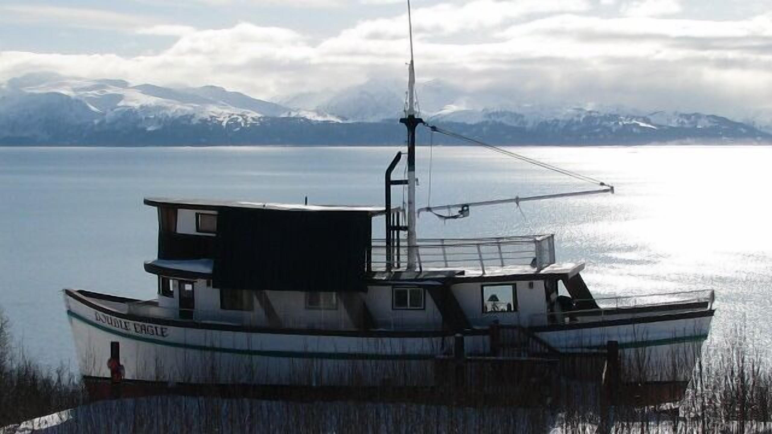 A boat with a cabin and a small deck area, docked near a body of water with mountains in the background.