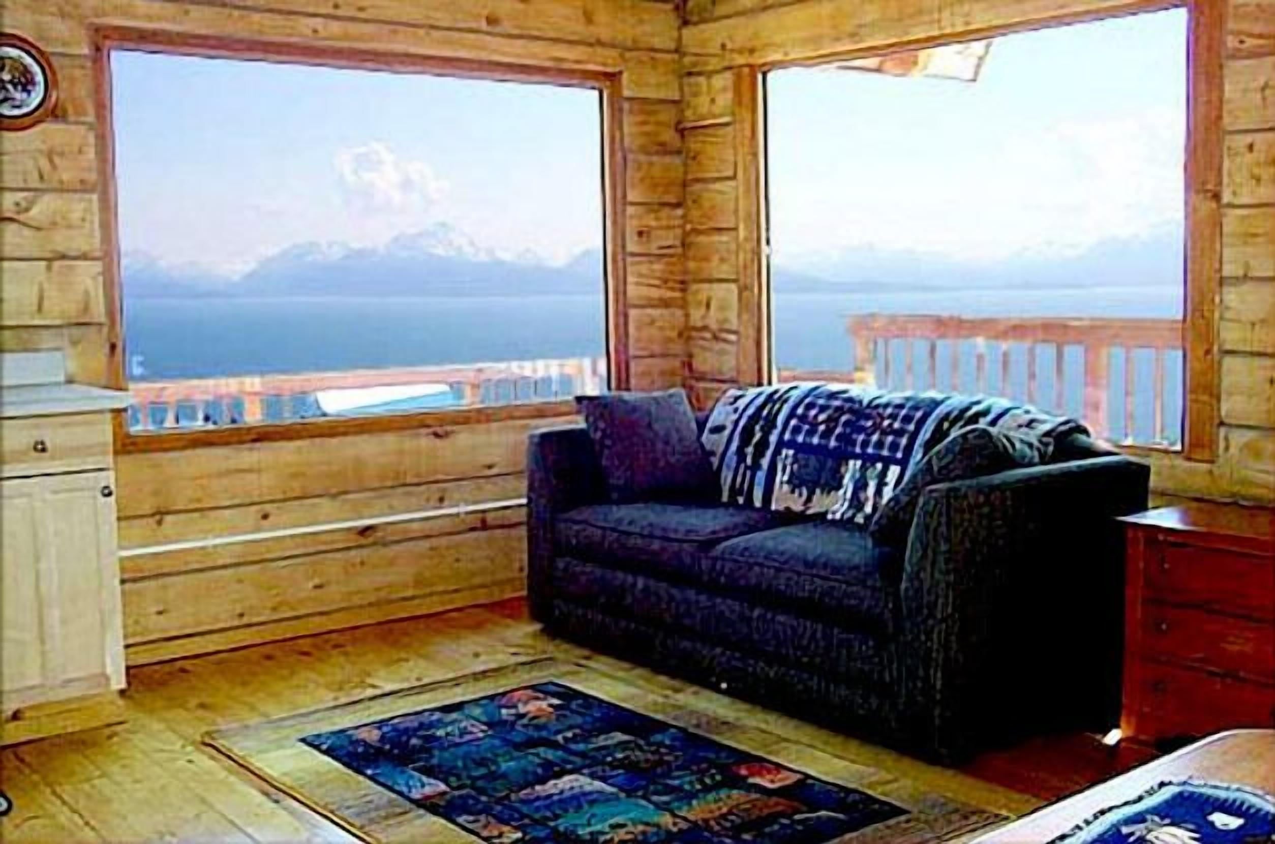 A wooden cabin room with a sofa and a rug, large windows with a view of a lake and mountains.