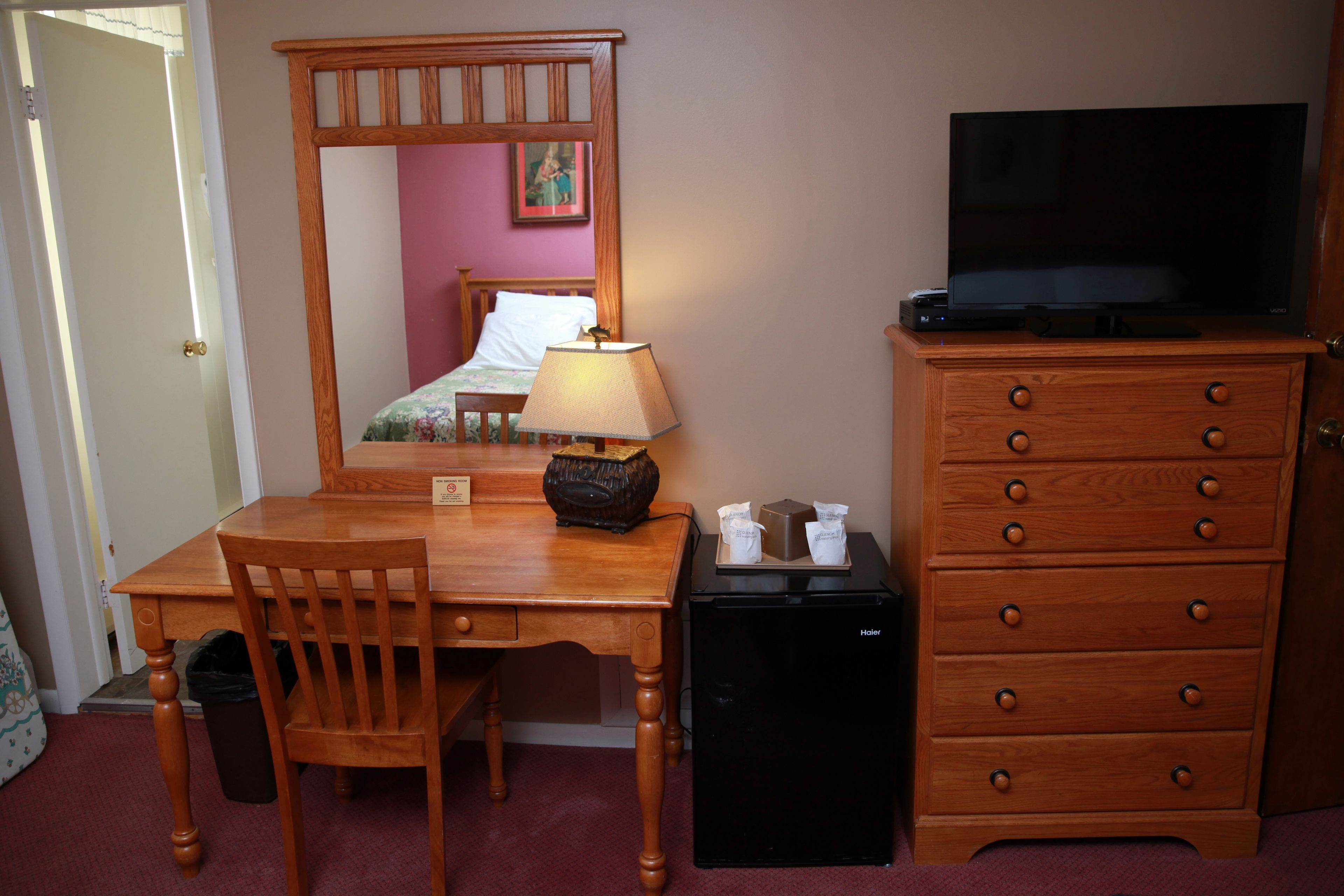 A hotel room with a wooden desk, a chair, a dresser, a television, and a small refrigerator.