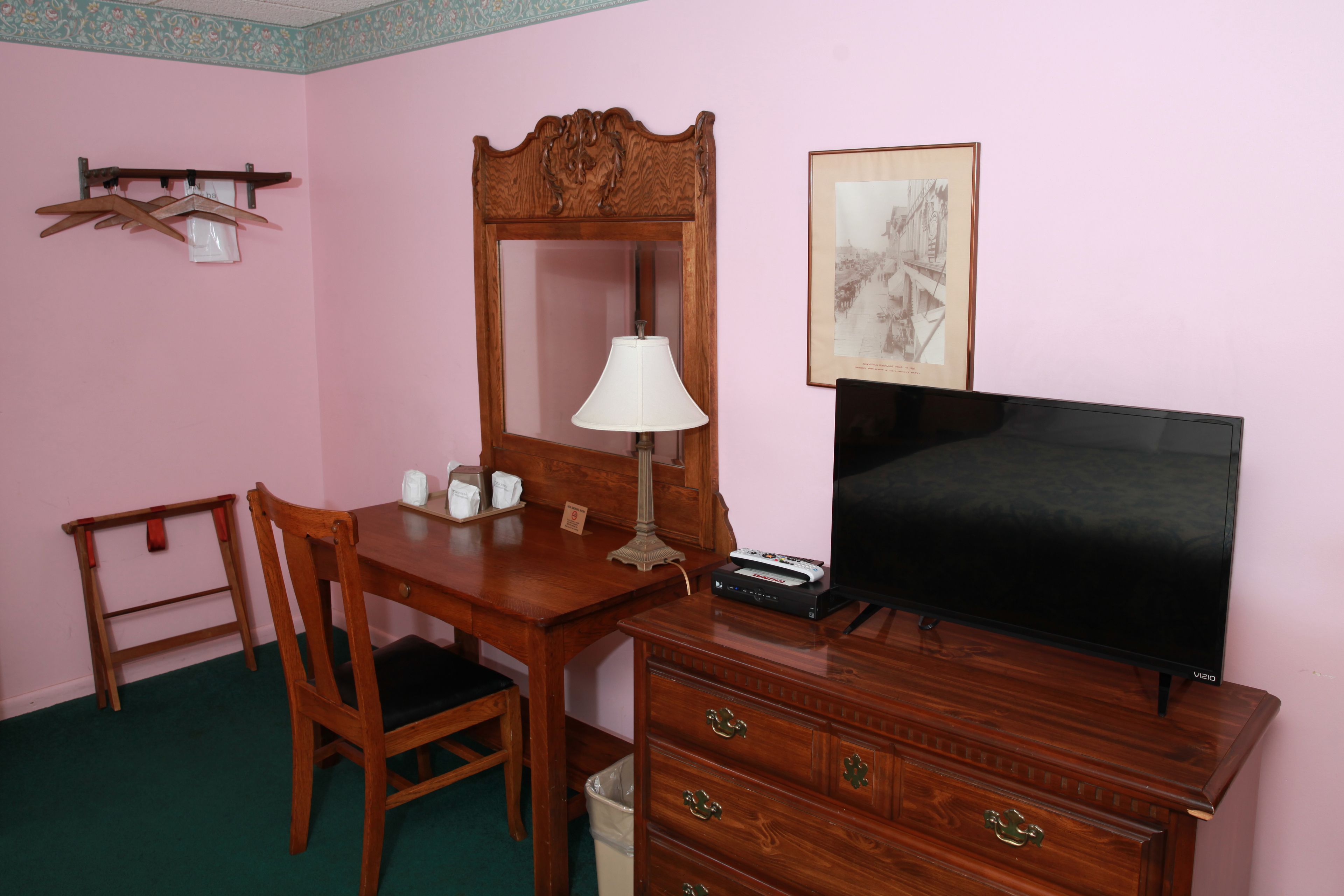 Una habitación con escritorio de madera, una silla, televisor, lámpara y cómoda.