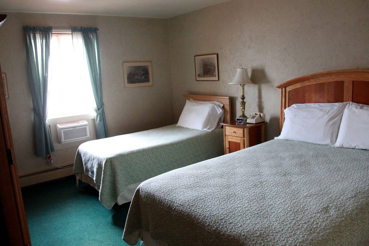 A hotel room with two single beds, a nightstand with a lamp, a telephone, and a window with curtains.