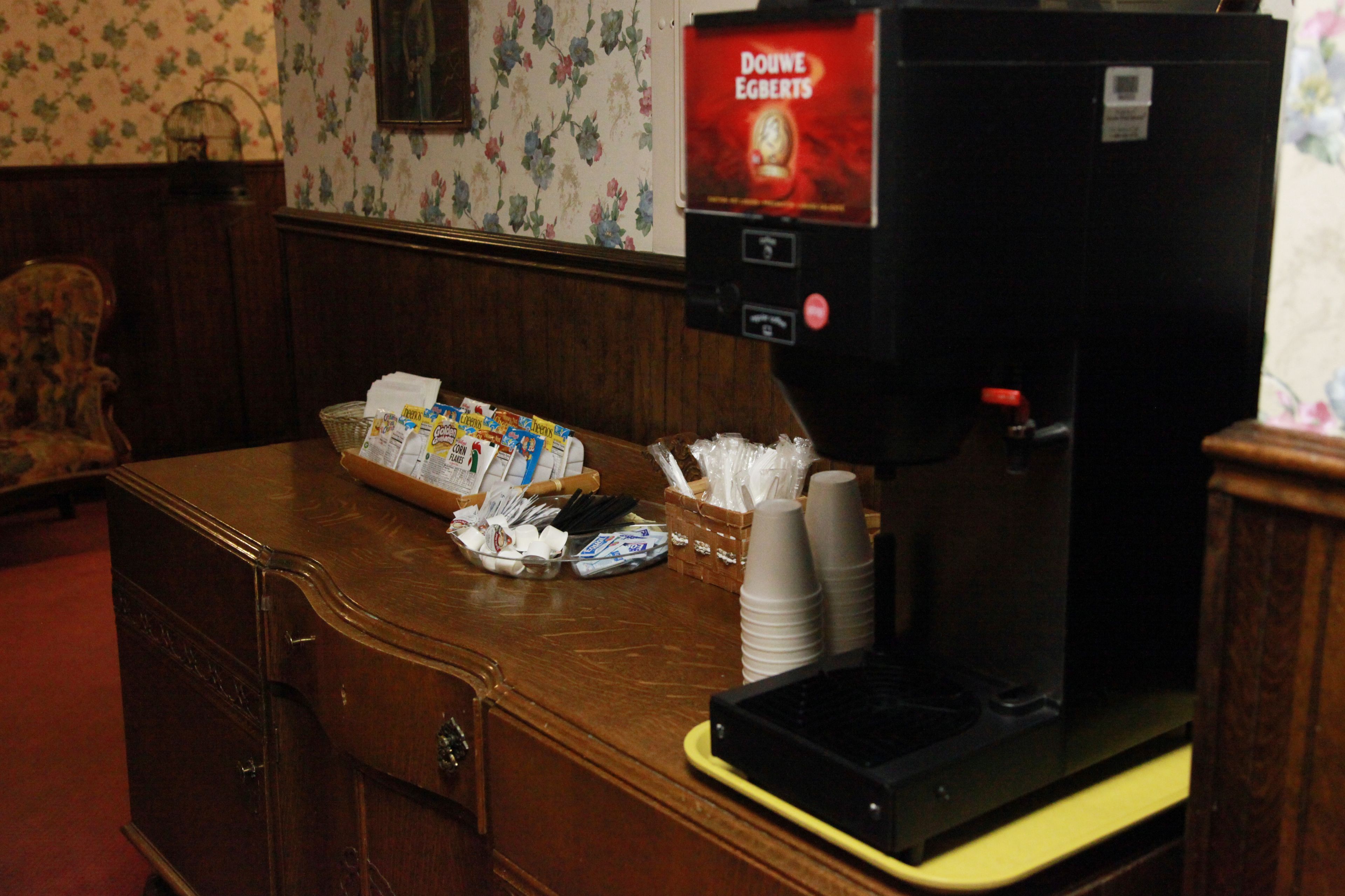 Una máquina de café con etiqueta Douwe Egberts, una cesta de aperitivos y vasos de papel apilados sobre una encimera de madera.