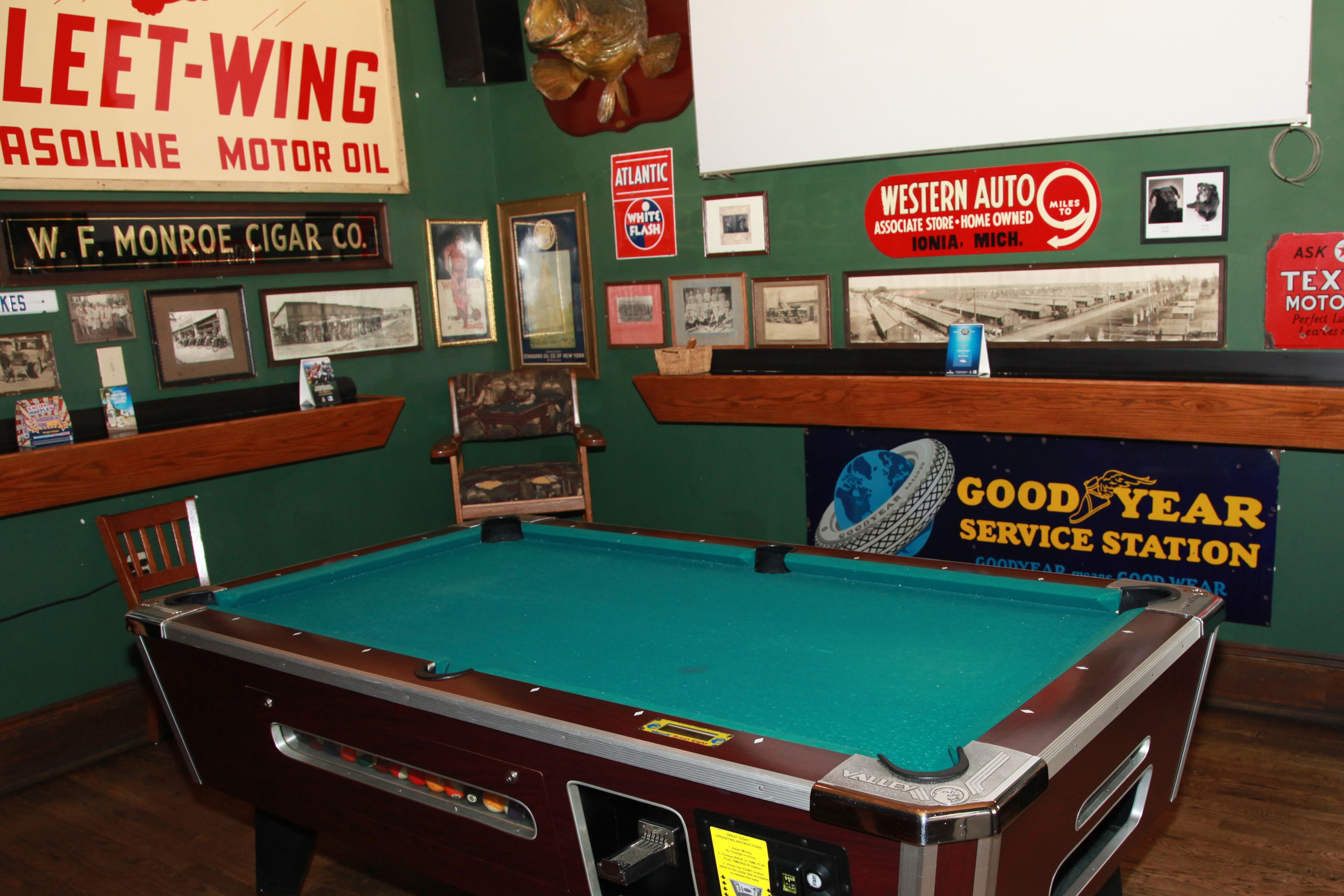 A pool table in a room with green walls, surrounded by various framed pictures and signs, including one for 'Goodyear Service Station'.