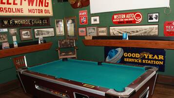 A pool table in a room with green walls, surrounded by various framed pictures and signs, including one for 'Goodyear Service Station'.
