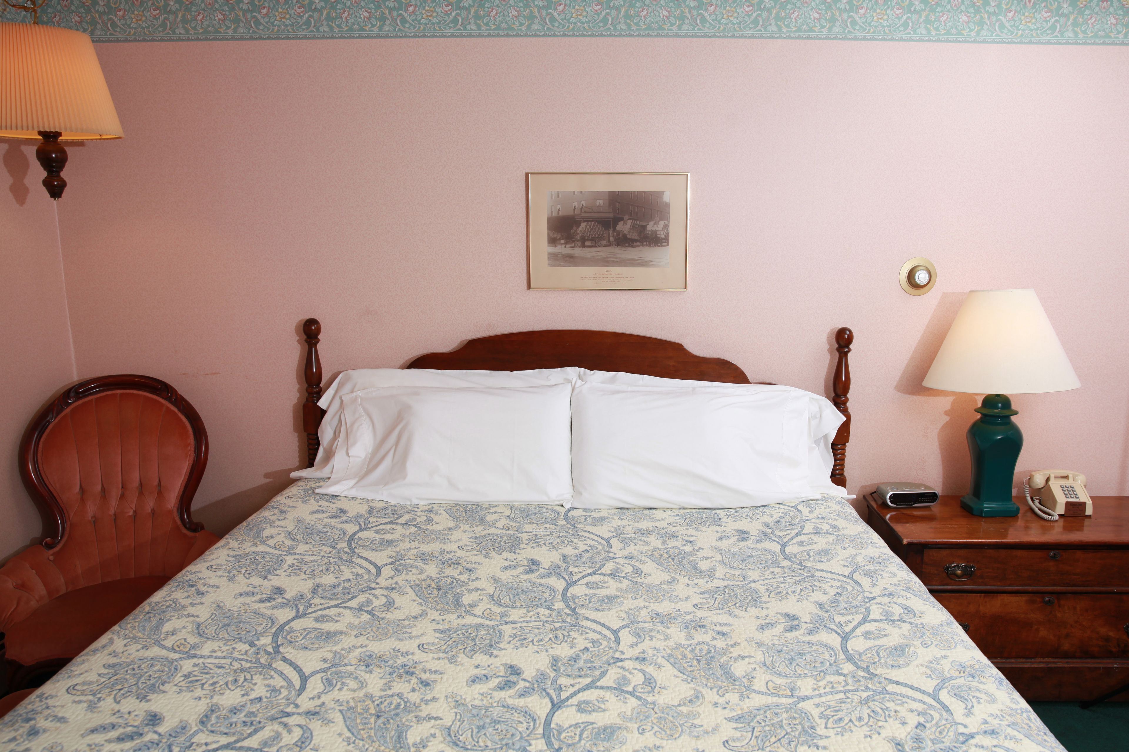 A bedroom with a bed, a wooden chair, a nightstand with a lamp, and a framed picture on the wall.