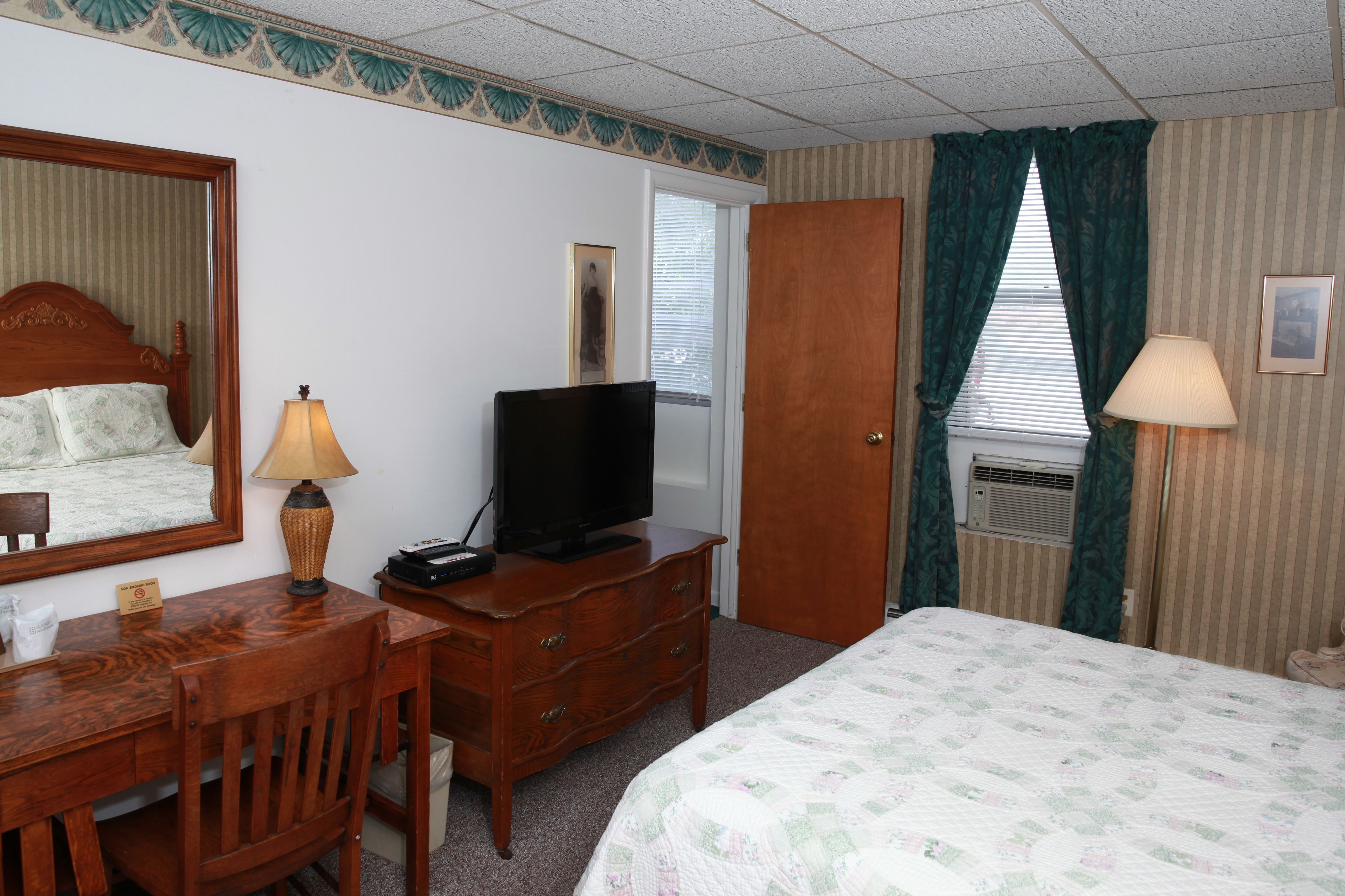 Habitación de hotel con cama, cómoda de madera, televisor, escritorio con lámpara y espejo.