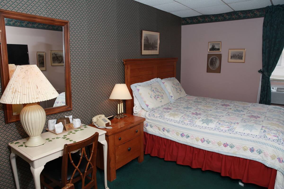 A hotel room with a wooden bed, a desk with a lamp, a chair, and a mirror. There are framed pictures on the wall and a window with curtains.