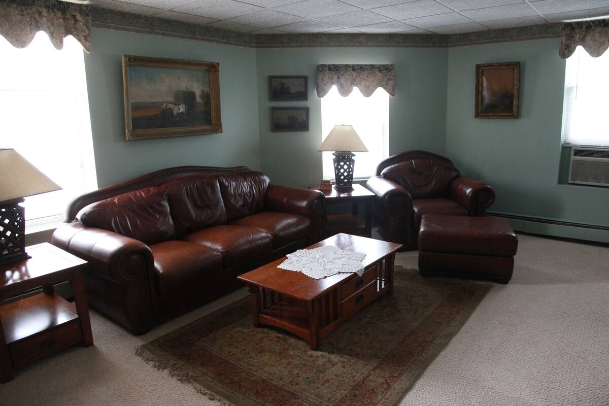 A living room with a brown leather sofa, a wooden coffee table, and a patterned rug. There are two framed pictures on the wall, a lamp on a side table, and a window with a curtain.