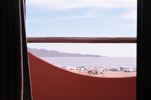 Vista de una playa a través de una ventana con marco de madera, montañas al fondo y gente y sombrillas en la playa.
