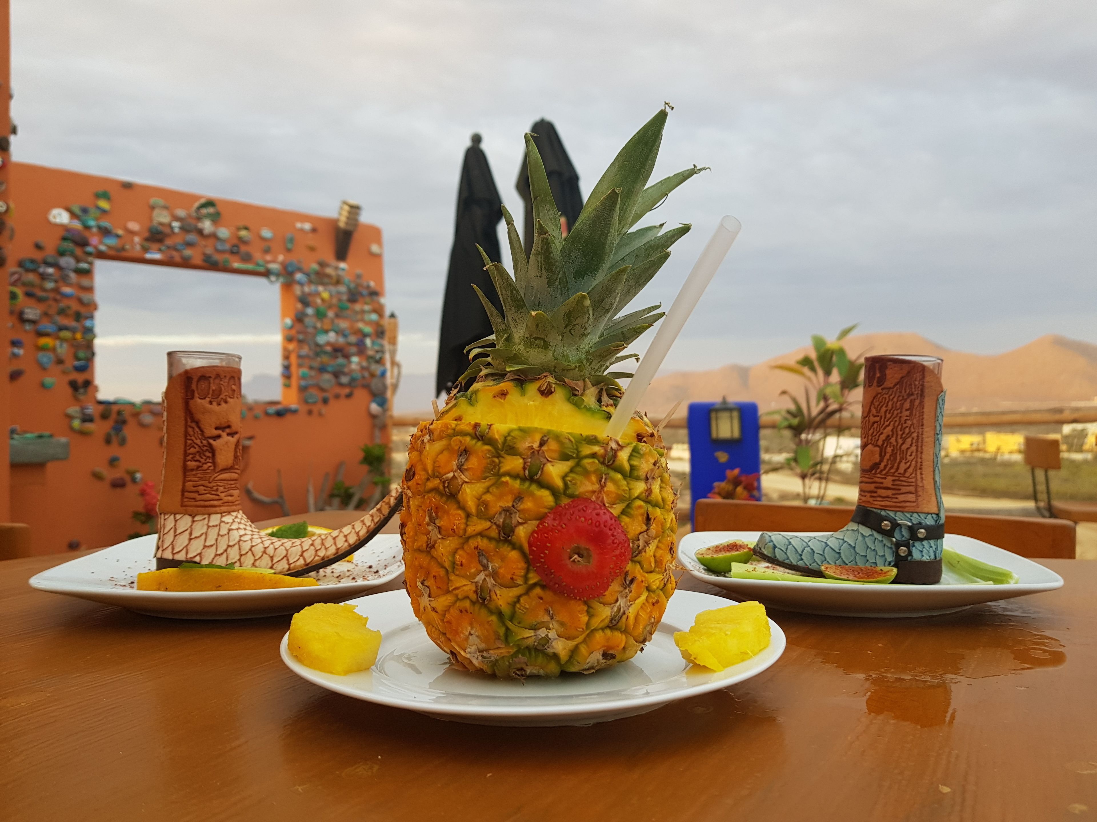 A pineapple with a straw and a strawberry on a plate, accompanied by a slice of yellow fruit and a small dish of green fruit, set on a table outdoors with a decorative wall in the background.