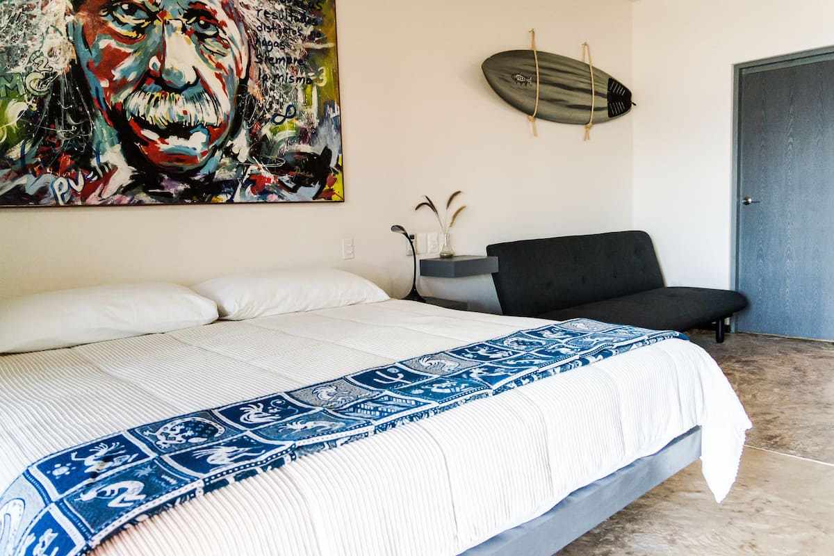 Une chambre avec un grand lit, une chaise noire, une planche de surf et un tableau coloré accroché au mur.