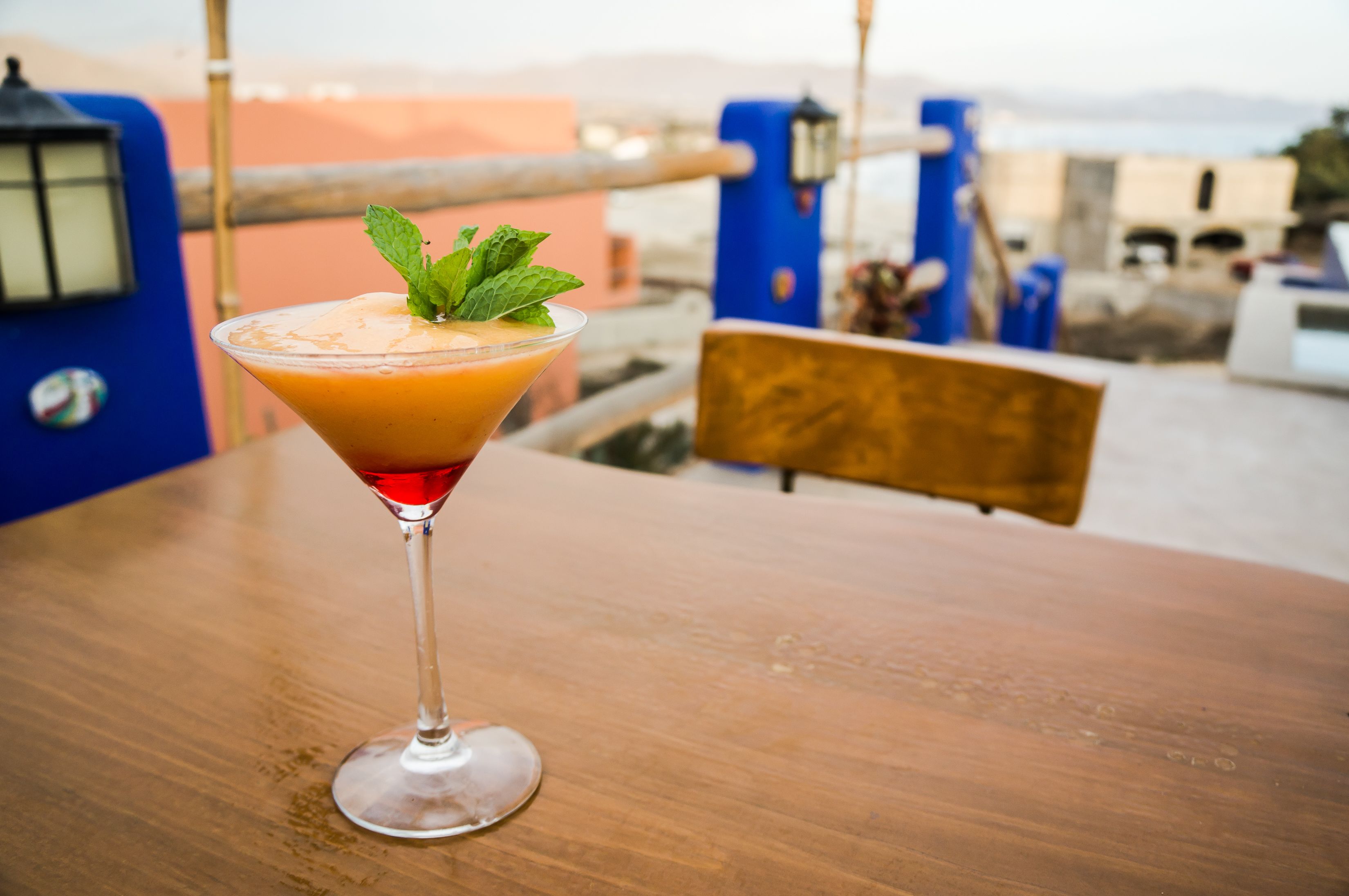 A martini glass with a garnish on a wooden table, set against a backdrop of a cityscape with buildings and a clear sky.