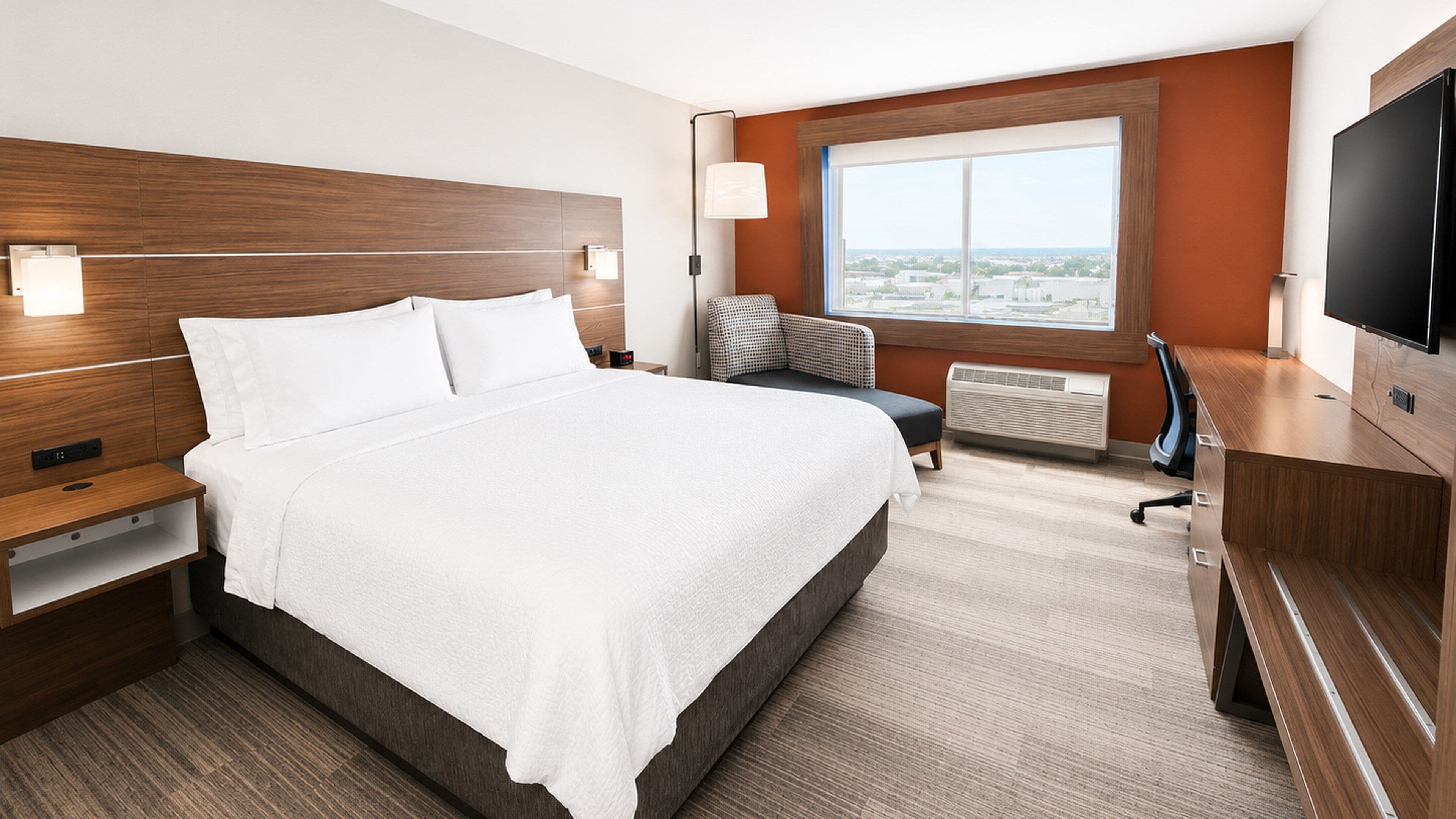 A hotel room with a large bed, a desk, a chair, a TV, and a window with a city view.