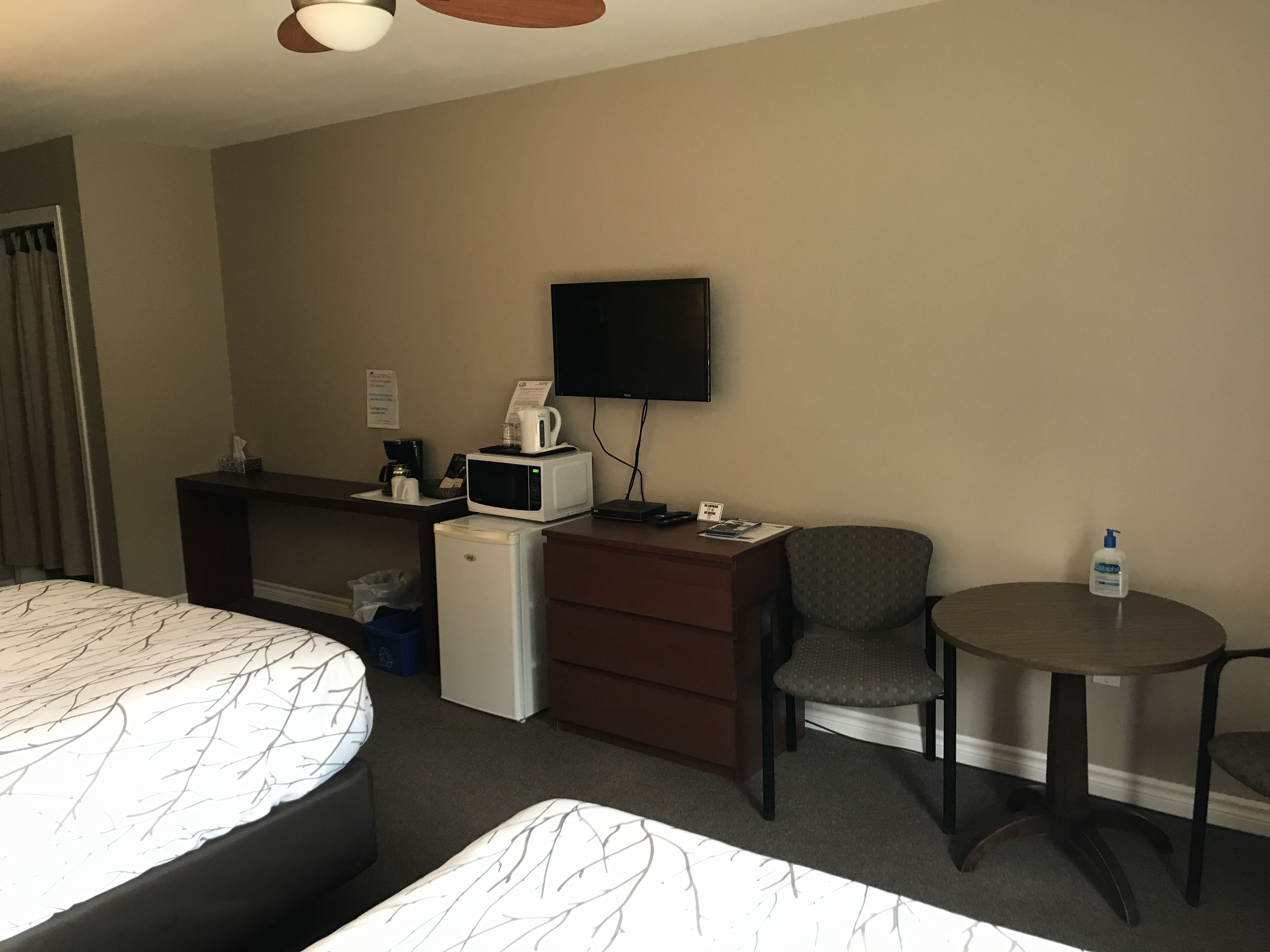A hotel room with a bed, a desk with a microwave, a chair, a small table, and a television on the wall.