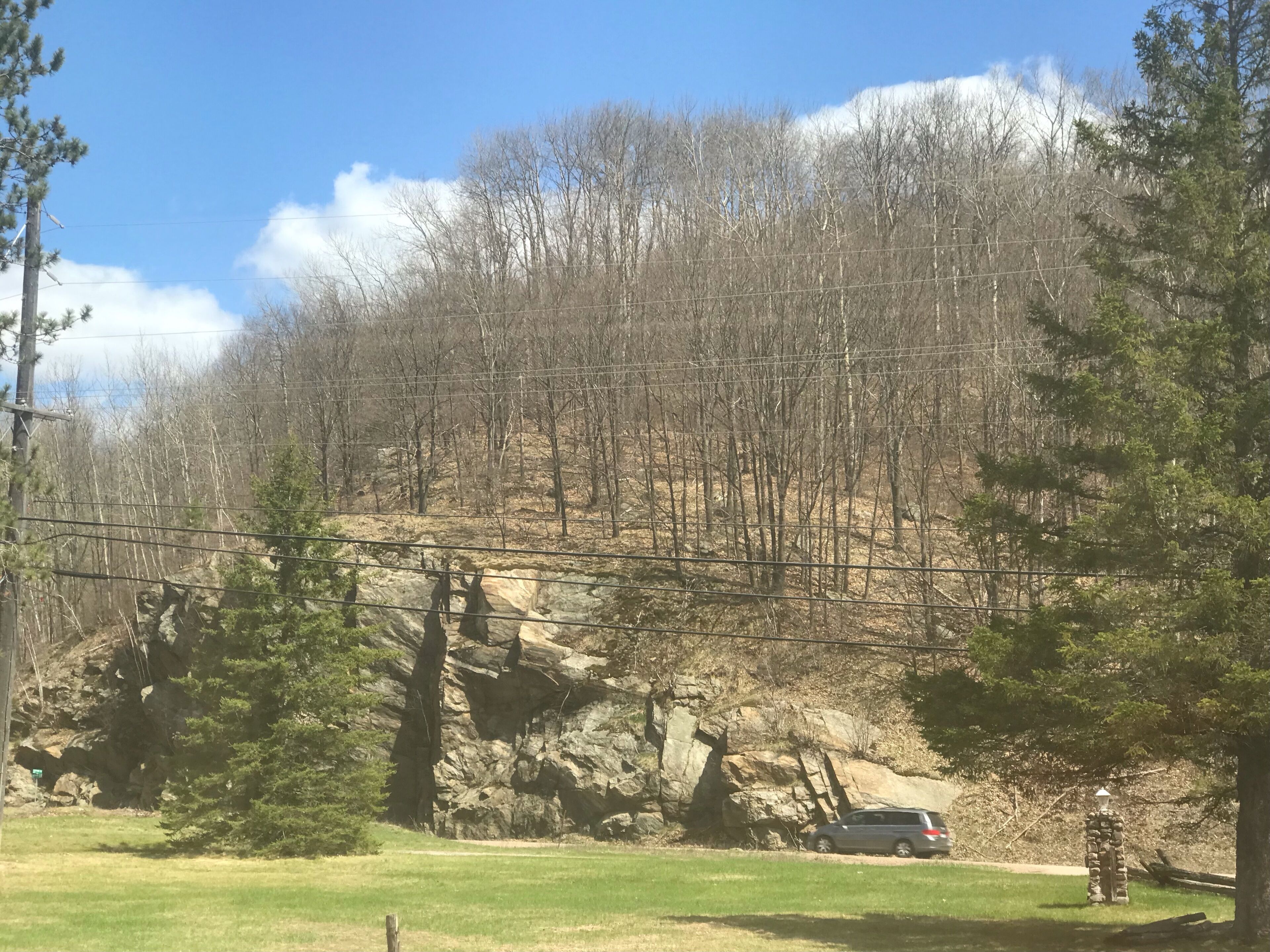 A natural landscape with a rocky outcrop, trees, and a car parked on a grassy area.