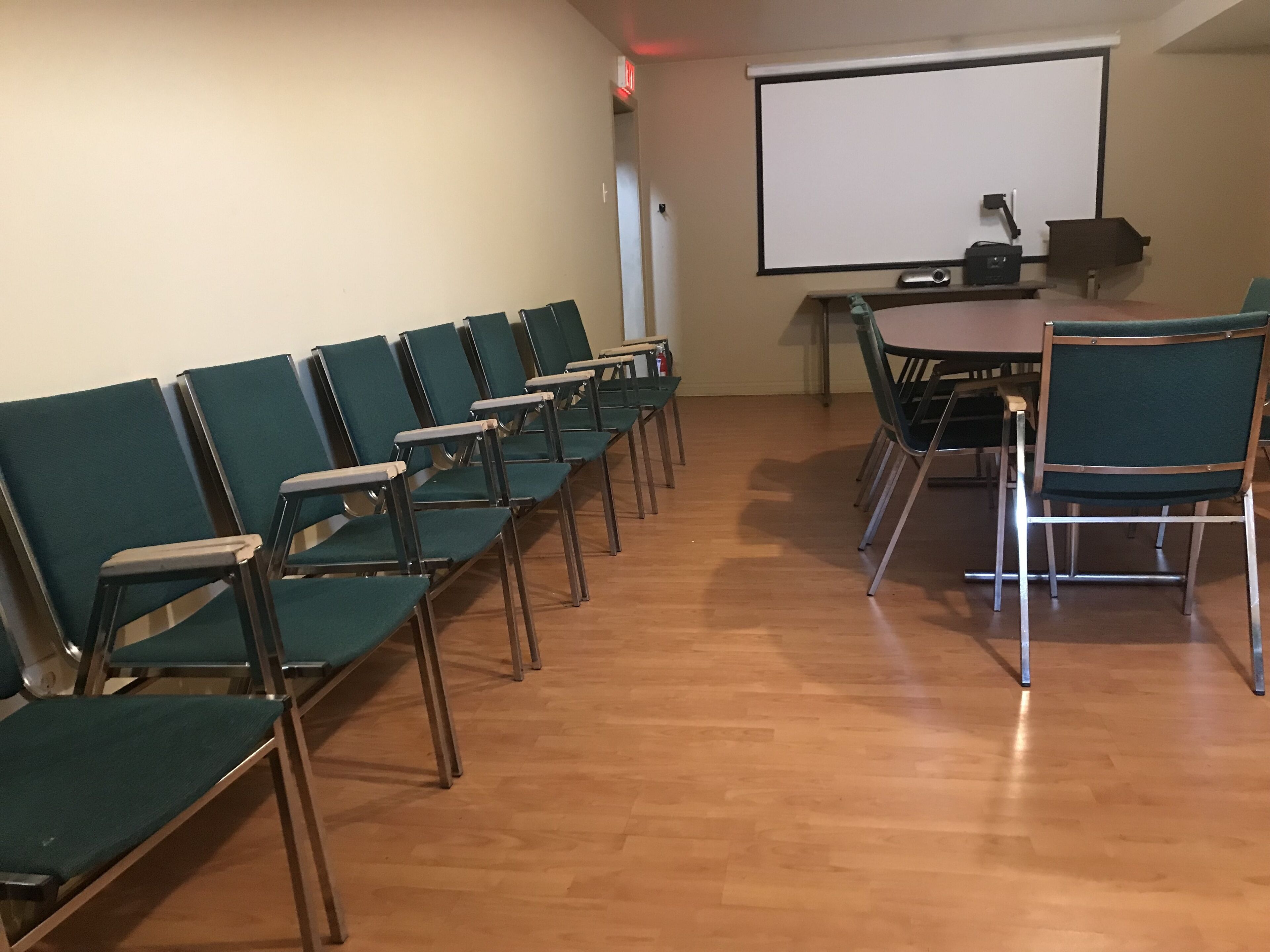 Un salón de conferencias con filas de sillas verdes, suelo de madera, una pantalla de proyector y una mesa con sillas.