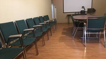 Un salón de conferencias con filas de sillas verdes, suelo de madera, una pantalla de proyector y una mesa con sillas.