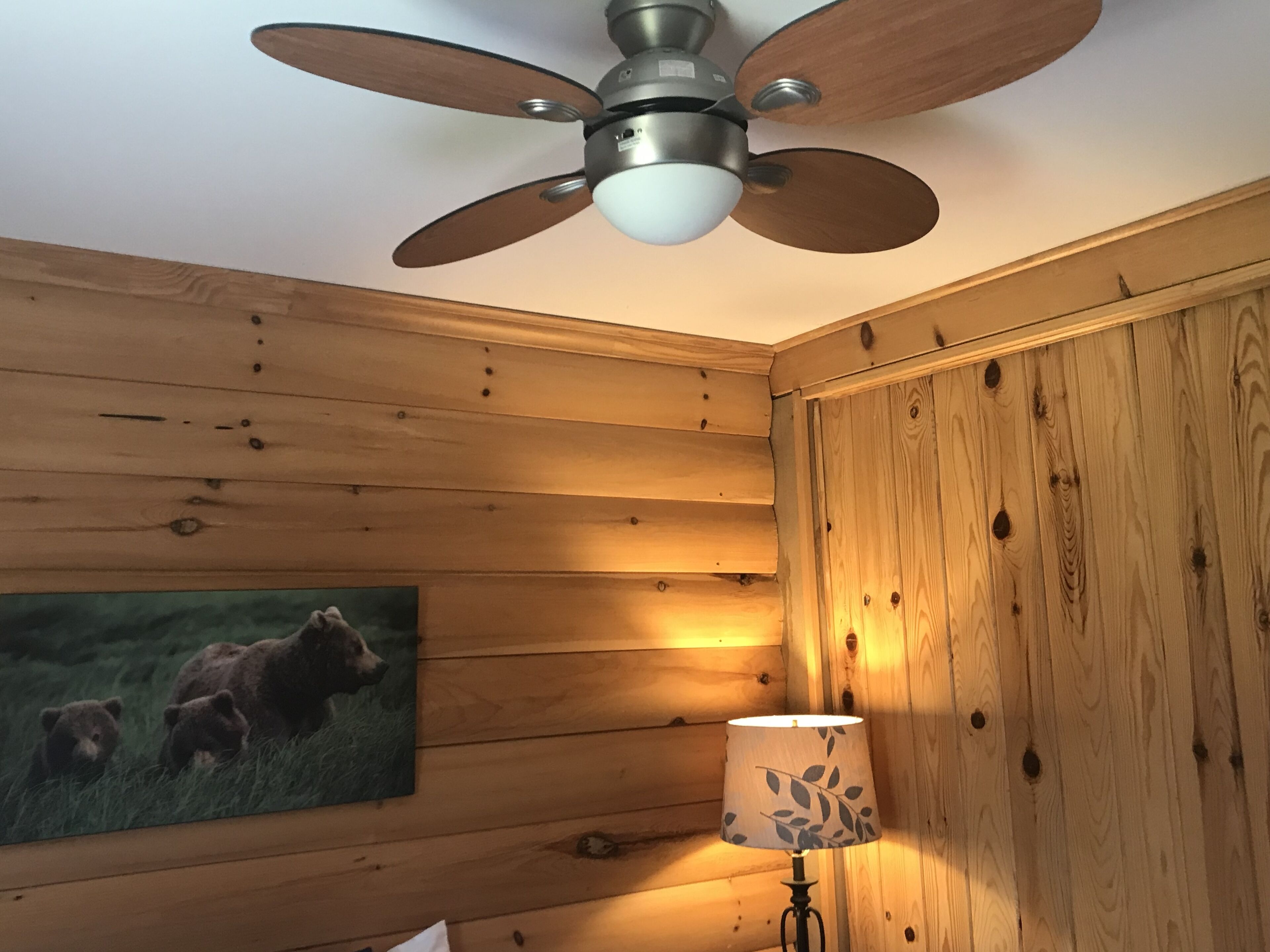 A room with wooden walls, a ceiling fan, a lamp, and a painting of bears.
