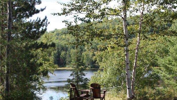 Un entorno junto al lago con una mesa de madera y dos sillas Adirondack mirando a un lago tranquilo, rodeado de un bosque denso.