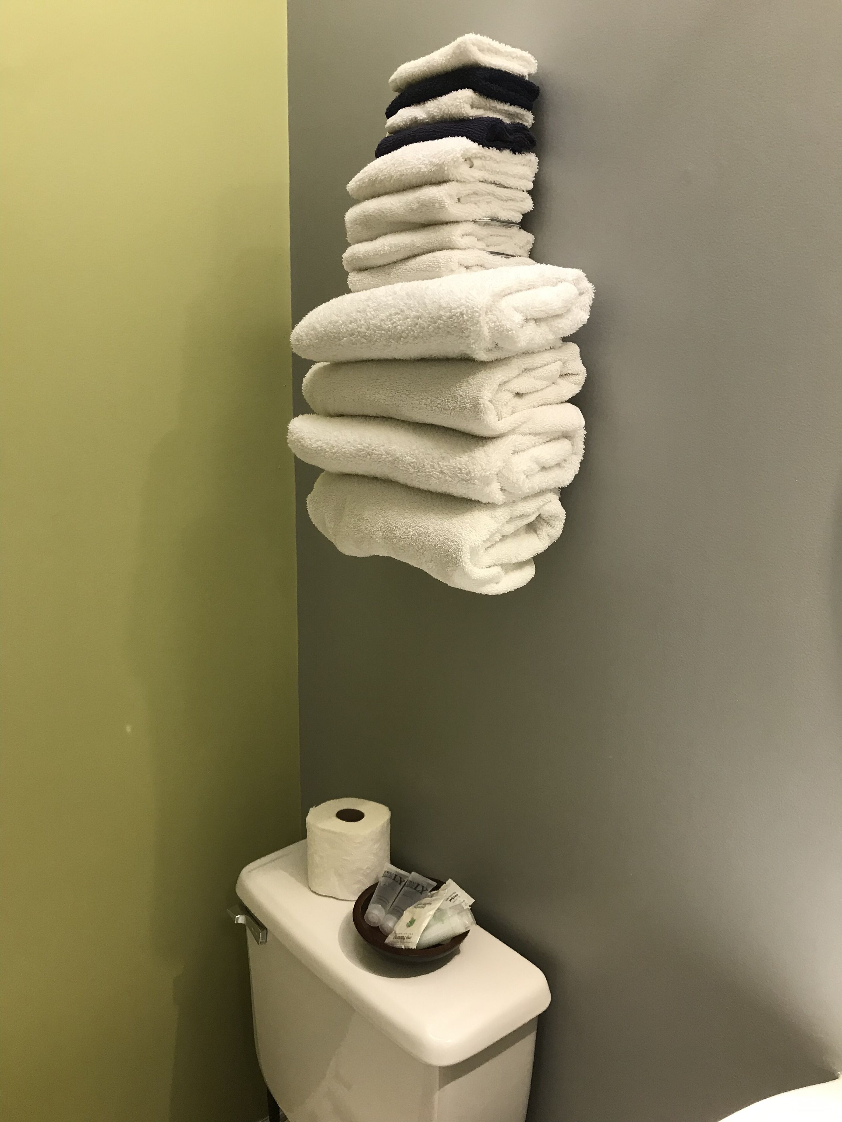 A stack of white towels hanging on a wall next to a toilet with a roll of toilet paper and a small bowl with toiletries.