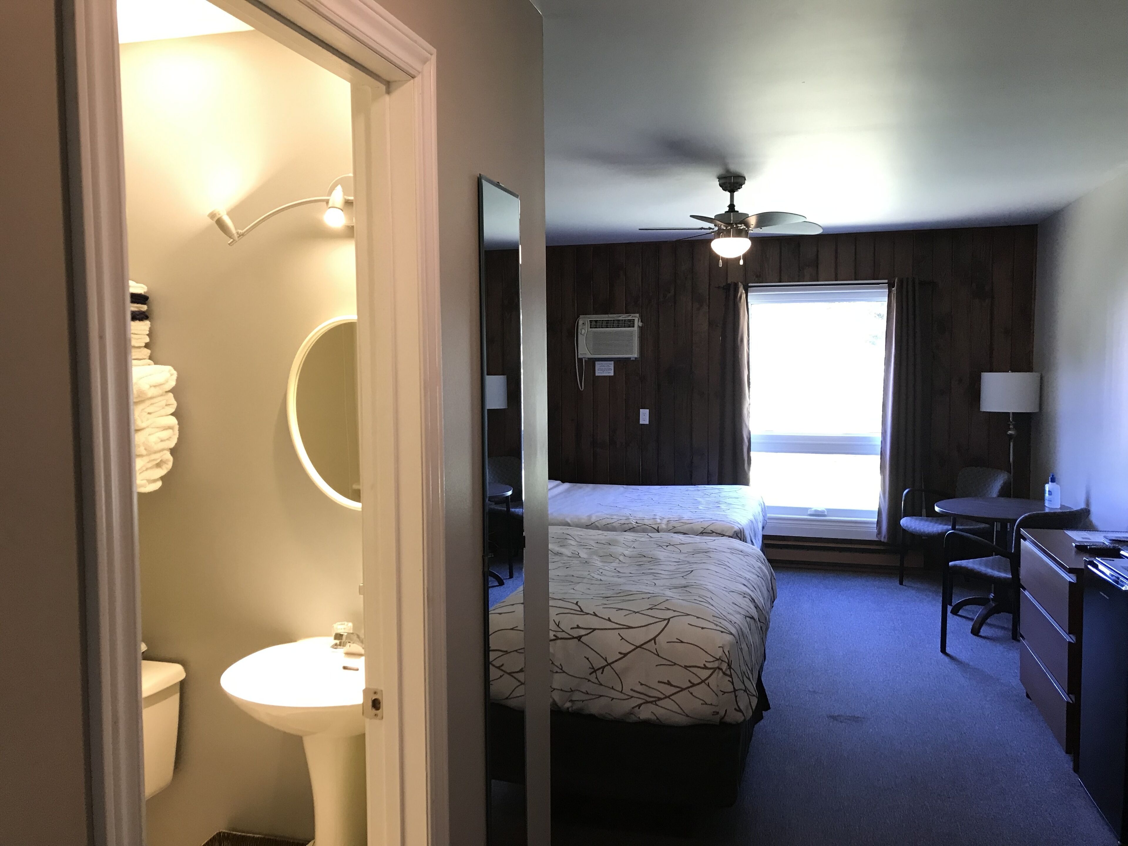 A hotel room with a bed, a desk, a chair, a ceiling fan, a window, and a bathroom with a toilet and sink.