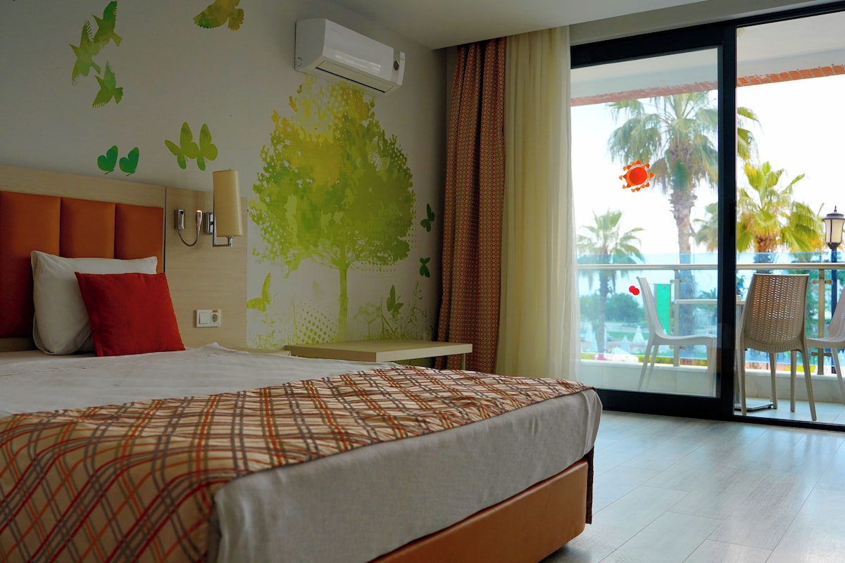 A hotel room with a bed, a nightstand, a wall-mounted air conditioner, a window with a view of palm trees, and a balcony with chairs.