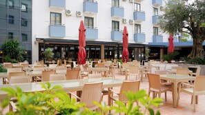 An outdoor seating area with wicker tables and chairs, red umbrellas, and a building with balconies in the background. - Club Aqua Plaza (Alanya)