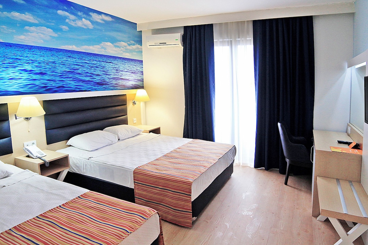 A hotel room with two beds, a desk, a chair, a TV, and a painting of the ocean.