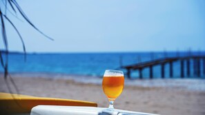 Ein Glas mit einem bernsteinfarbenen Getränk auf einer weißen Oberfläche, im Hintergrund ein Strand und ein Steg.