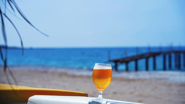 A glass of amber-colored beverage on a white surface with a beach and a pier in the background. - Club Aqua Plaza (Alanya)