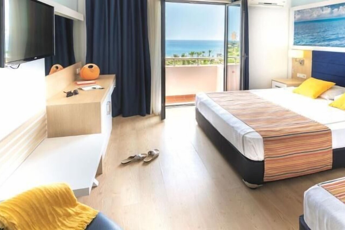 A hotel room with a bed, a desk, a TV, and a balcony with a view of the ocean.