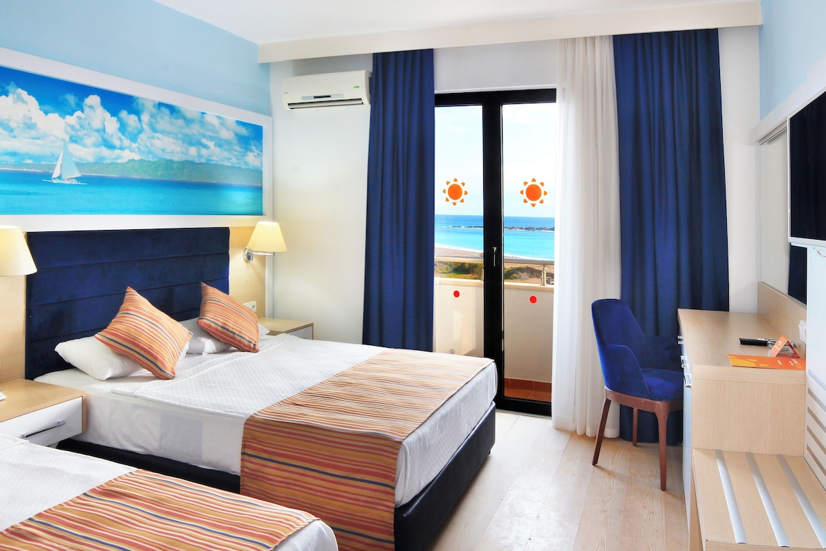 A hotel room with a bed, a desk, and a chair. There is a painting of a sailboat on the wall, and a window with curtains.