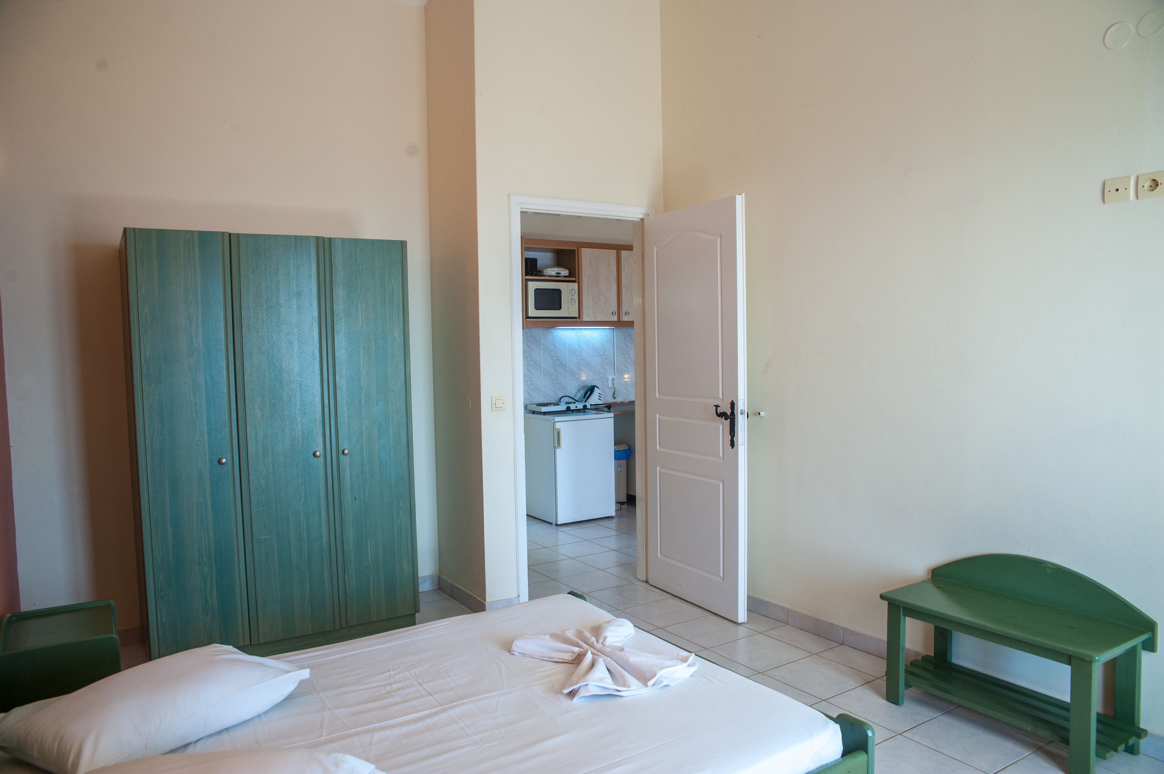 A bedroom with a bed, a green wardrobe, a green bench, and a view into a kitchen area.
