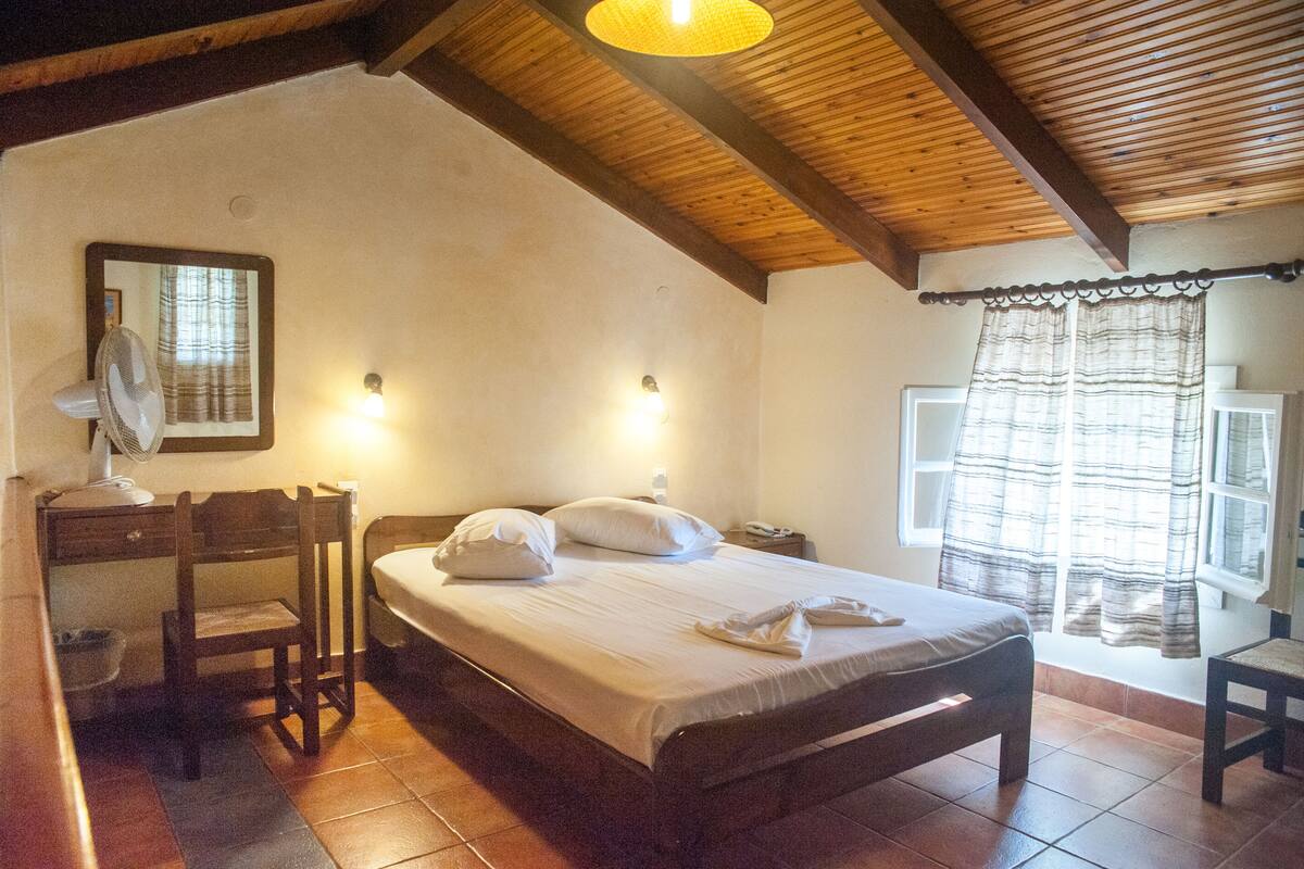 A bedroom with a wooden bed, a desk with a chair, a mirror, and a window with curtains.