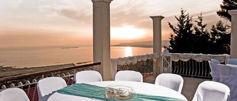 Une table ronde entourée de chaises blanches, agrémentée d’un chemin de table turquoise et d’un centre de table en verre, le tout mis en valeur par un décor de coucher de soleil avec des colonnes et des arbres en arrière-plan.