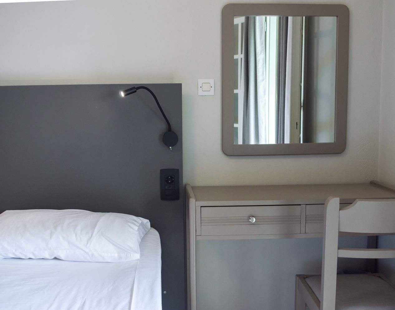 Habitación de hotel con cama, escritorio con cajones, espejo, lámpara de pared y panel de control.