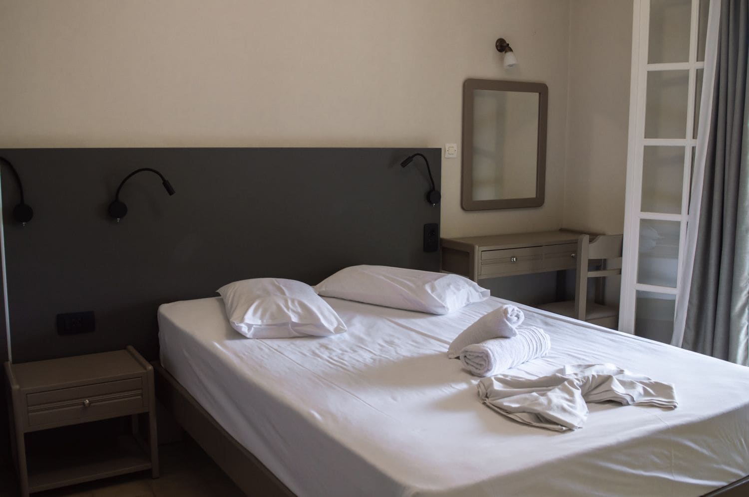A hotel room with a bed, a bedside table, a nightstand, a desk, a mirror, and a window with curtains.