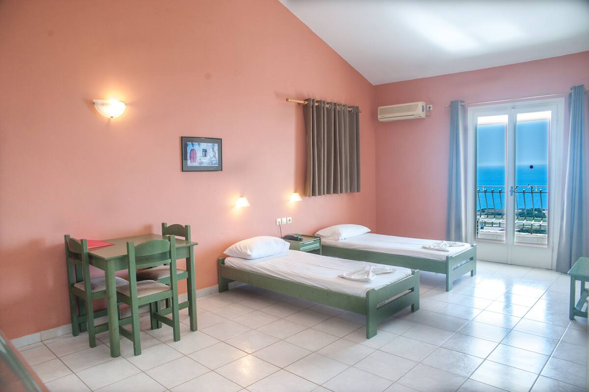 A room with two beds, a desk, and a chair. There is a window with curtains and a view of the sea.