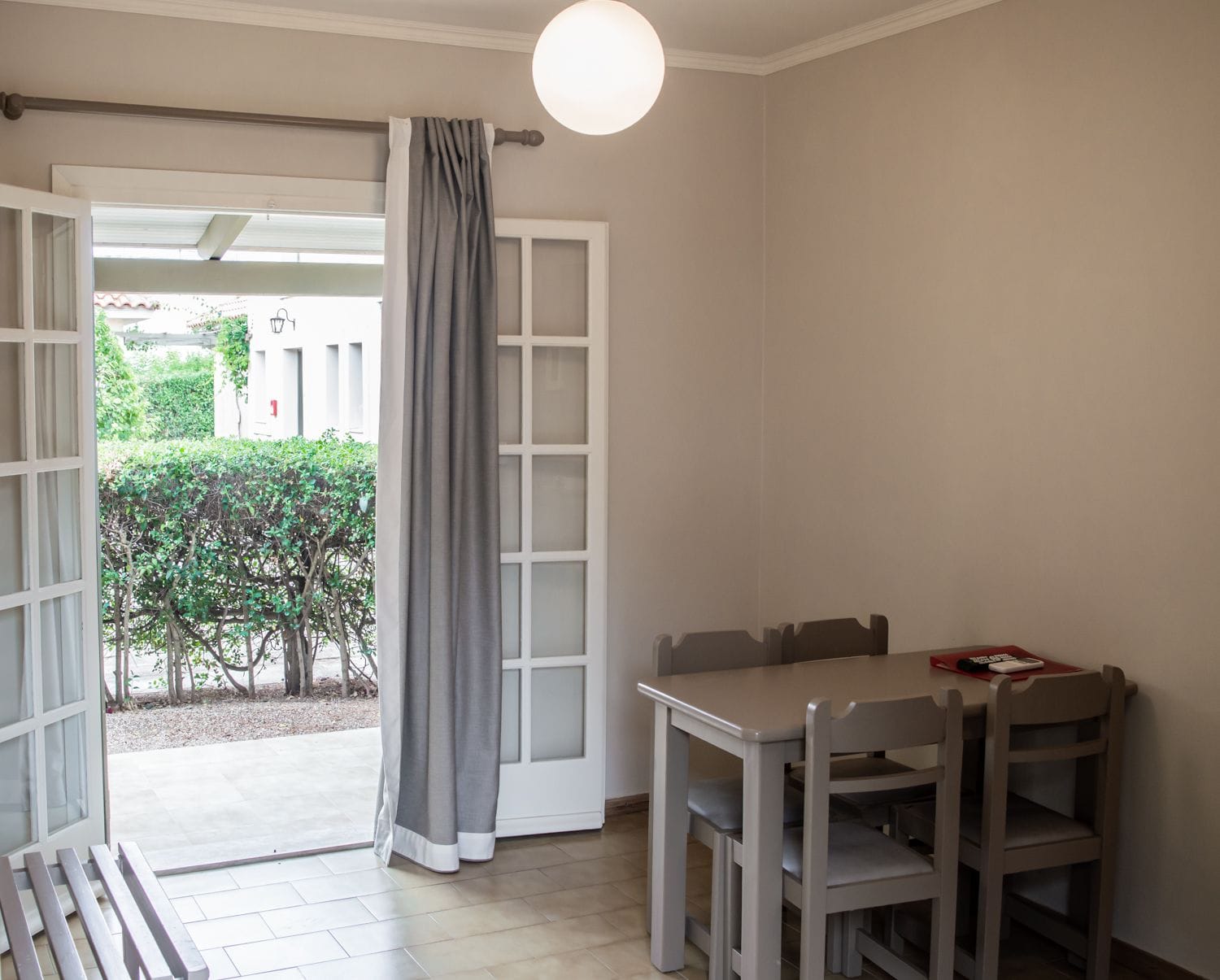 A room with a dining table and chairs, a window with a grey curtain, and a view of a garden and building outside.