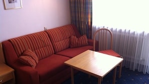 A red sofa with striped cushions, a wooden table, and a chair in a room with a blue carpet and a window with curtains. - Lifthotel (Kirchberg in Tirol)