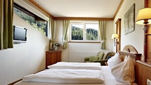 A hotel room with a bed, a desk, a chair, a window with curtains, a wall-mounted TV, and a lamp. - Lifthotel (Kirchberg in Tirol)