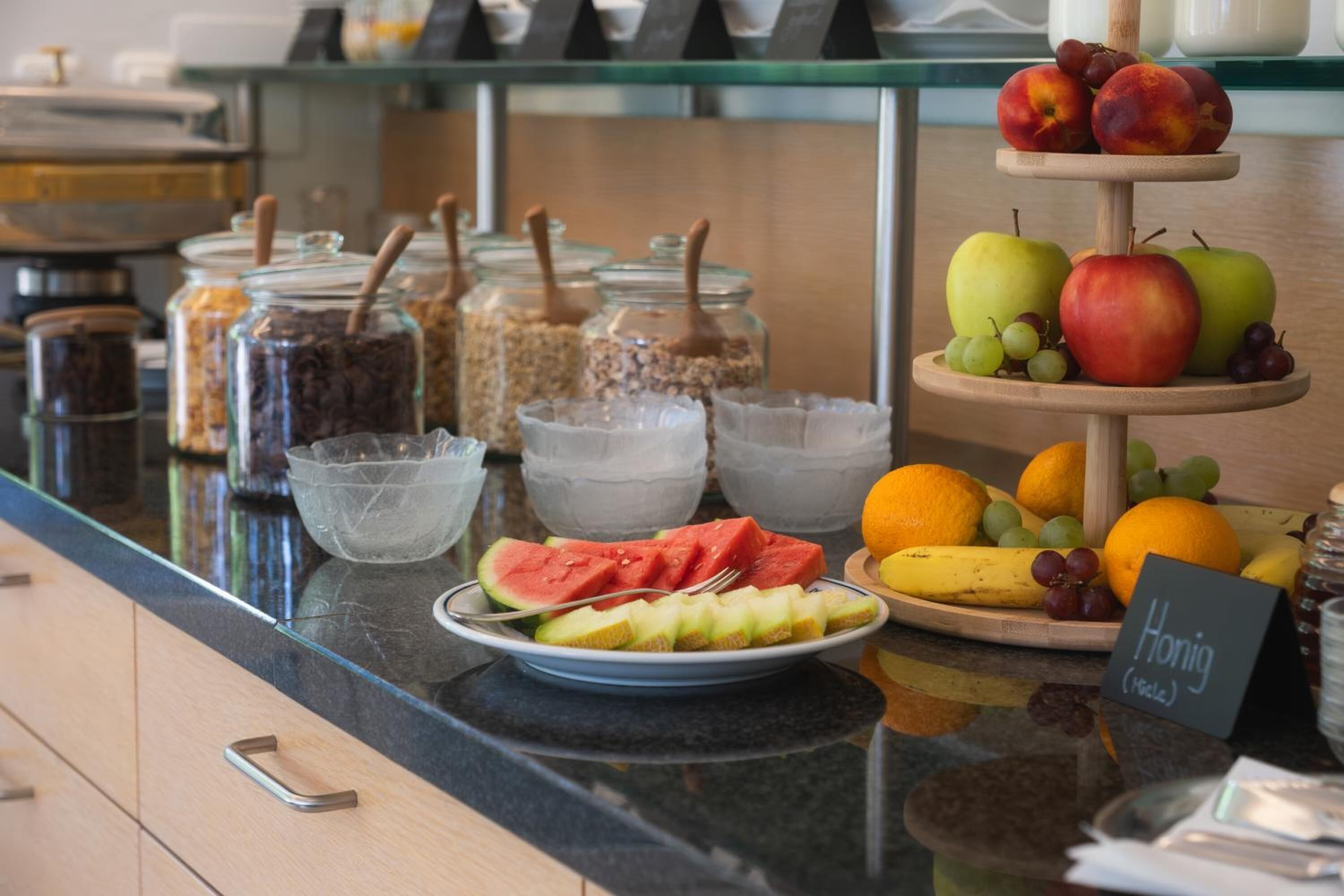 Un buffet de petit-déjeuner proposant divers fruits, une pancarte indiquant « melon miel », et des bocaux remplis de granola et d’autres produits pour le petit-déjeuner.