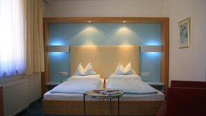 Cama doble con cabecera de madera, dos mesitas de noche, una mesita redonda y una silla roja en una habitación de hotel.