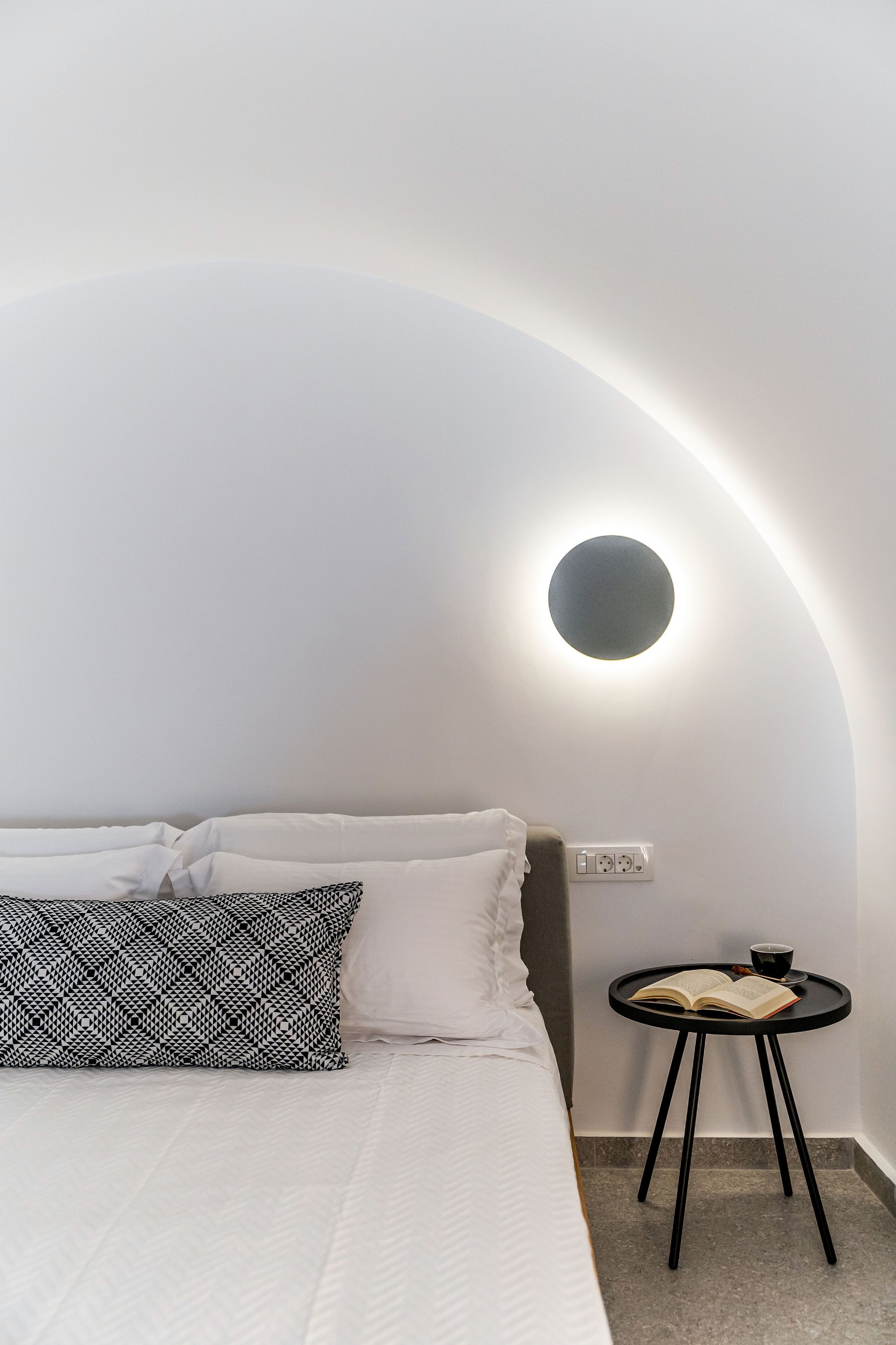 A modern bedroom with a bed, a bedside table with a book and a cup, and a wall-mounted light fixture.