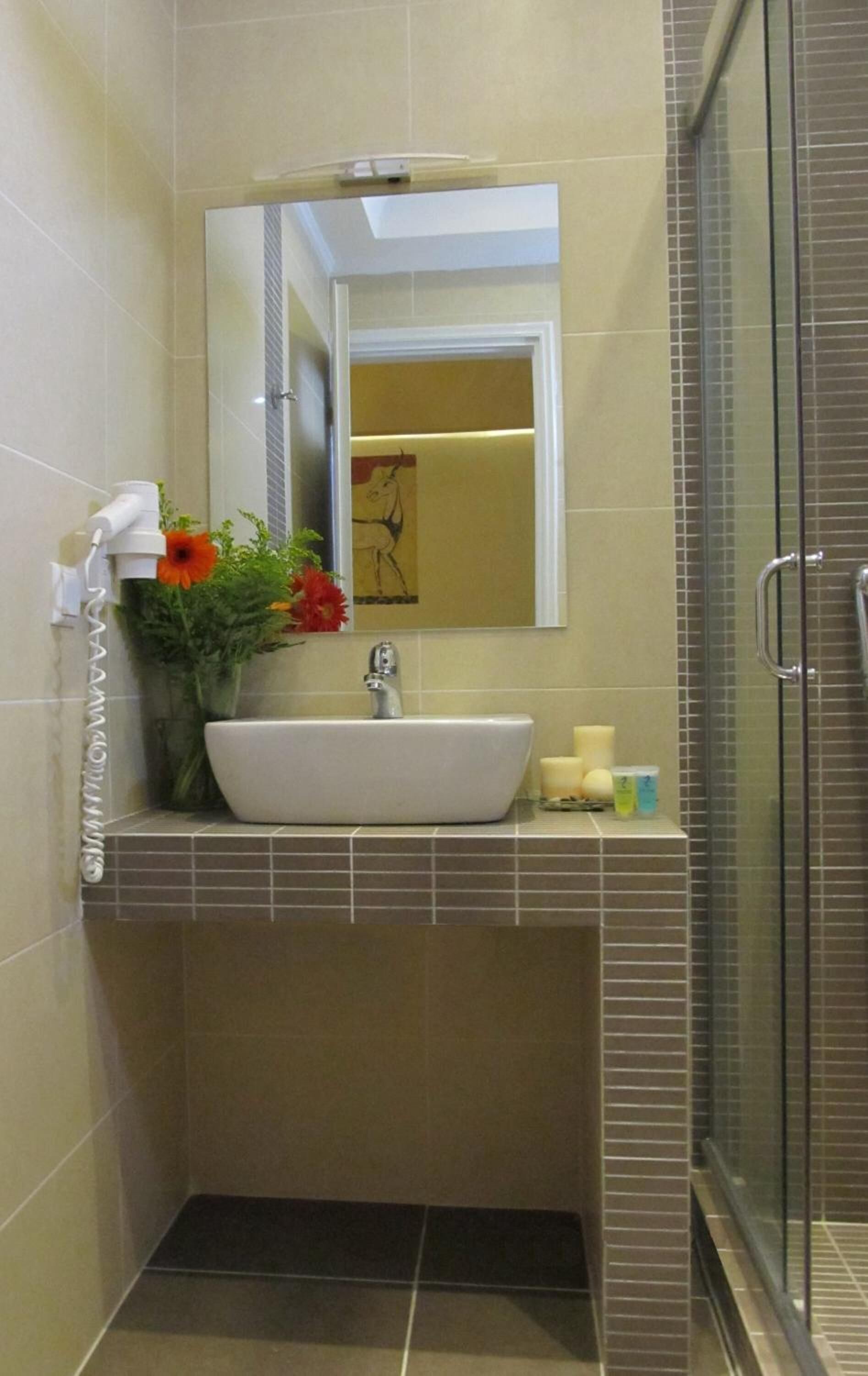 A bathroom with a white sink, a mirror, a shower, and a vase with flowers.