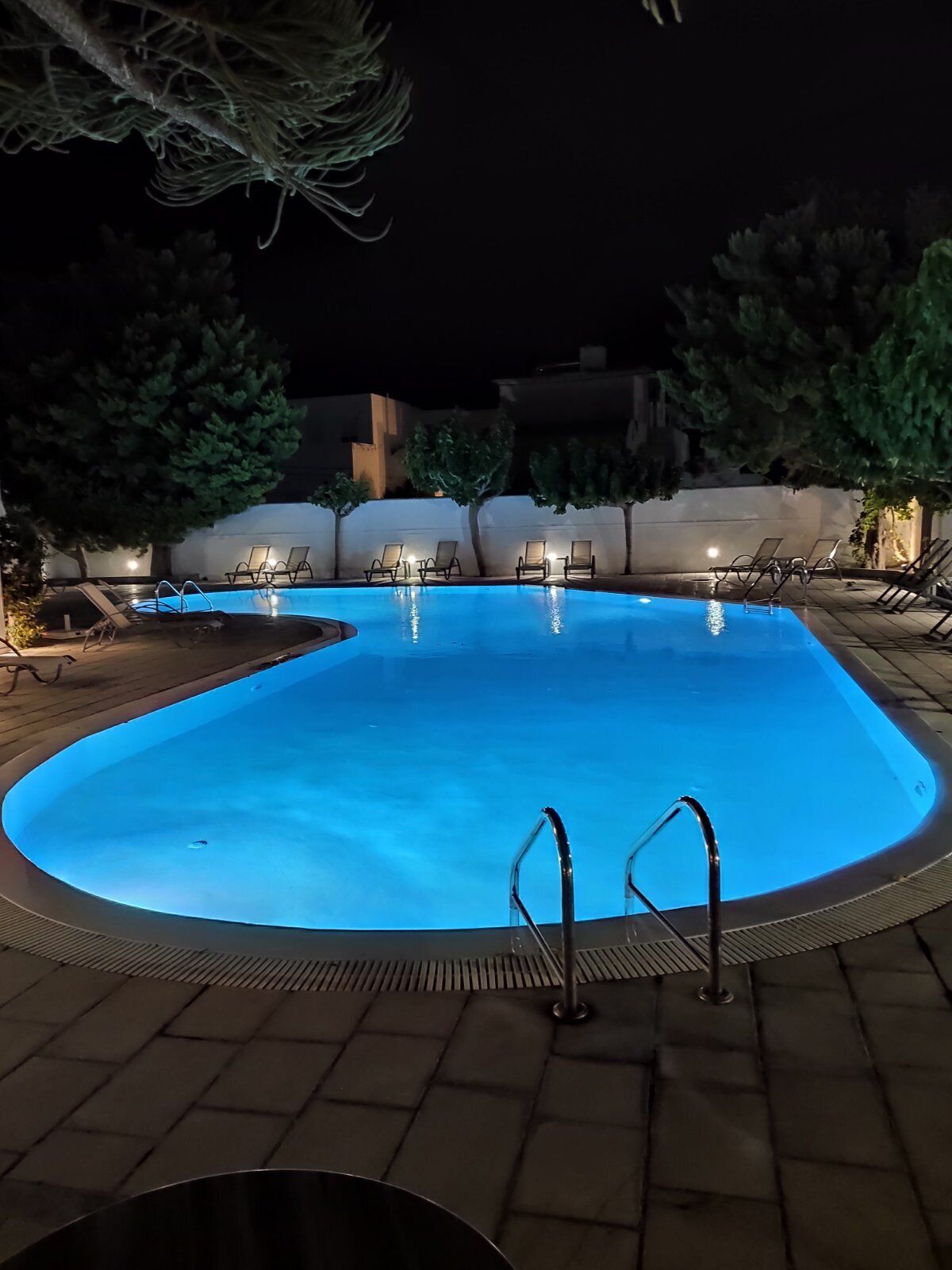 A nighttime swimming pool with clear water, surrounded by lounge chairs and a paved deck.