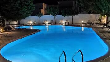 A nighttime swimming pool with clear water, surrounded by lounge chairs and a paved deck.