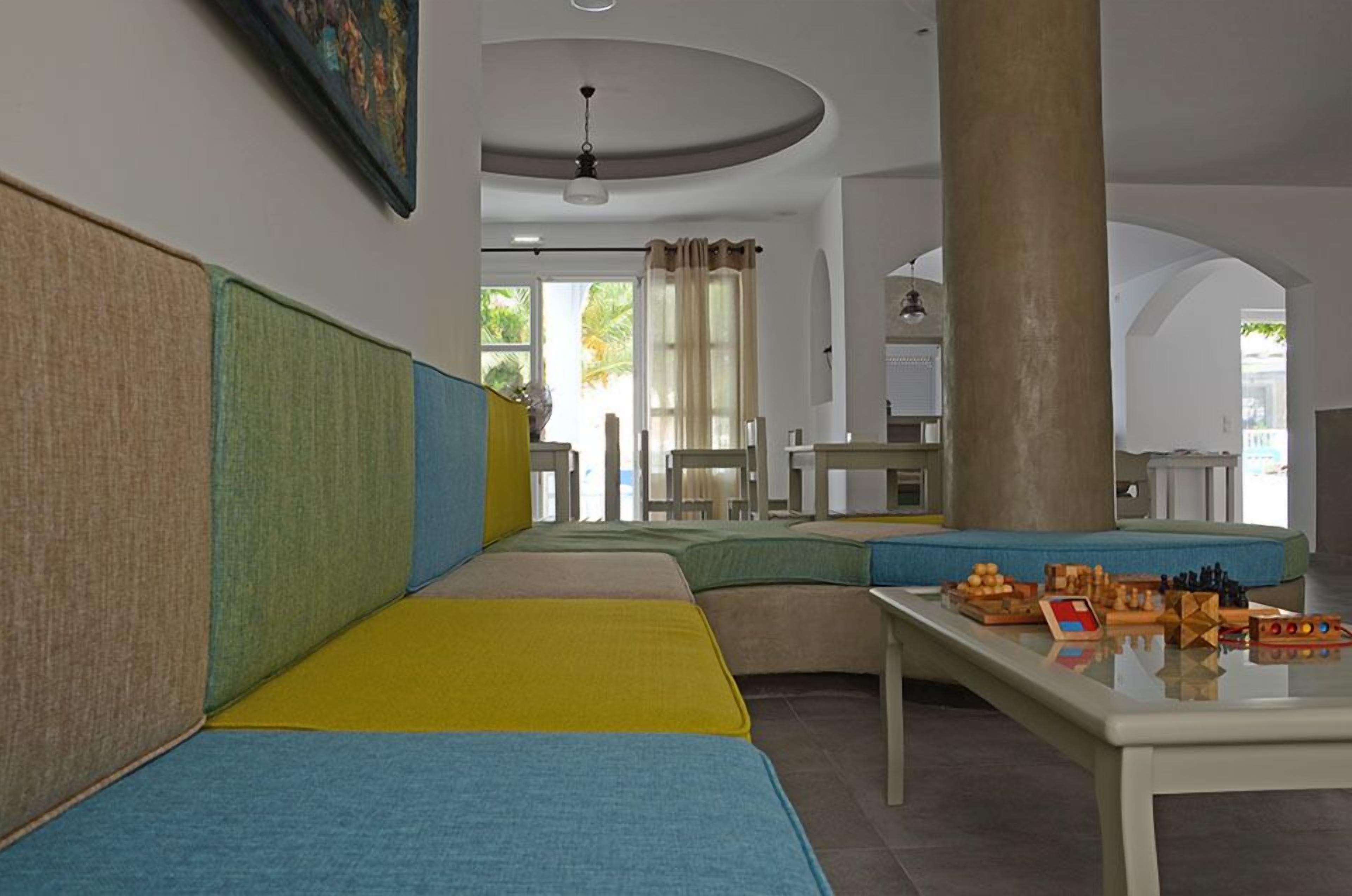 A hotel lobby with a long, curved seating area with colorful cushions, a central pillar, and a table with board games.